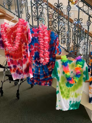 Three colorful tie-dye t-shirts hanging on hangers in front of a decorative wrought iron staircase railing. Each shirt is adorned with a vibrant flower lei. A walker is partially visible in the background on a carpeted floor.