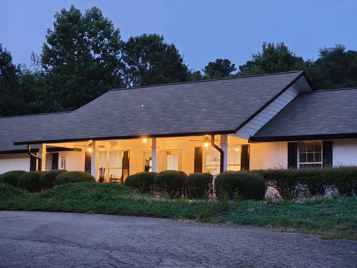 Single-story care facility front with lit entrance, manicured shrubs, and a driveway at dusk.