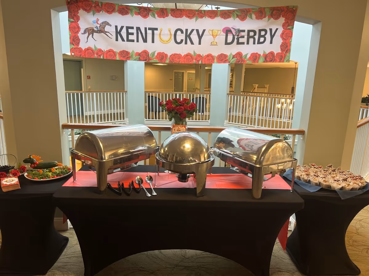 Buffet table with chafing dishes and desserts beneath a Kentucky Derby banner indoors.