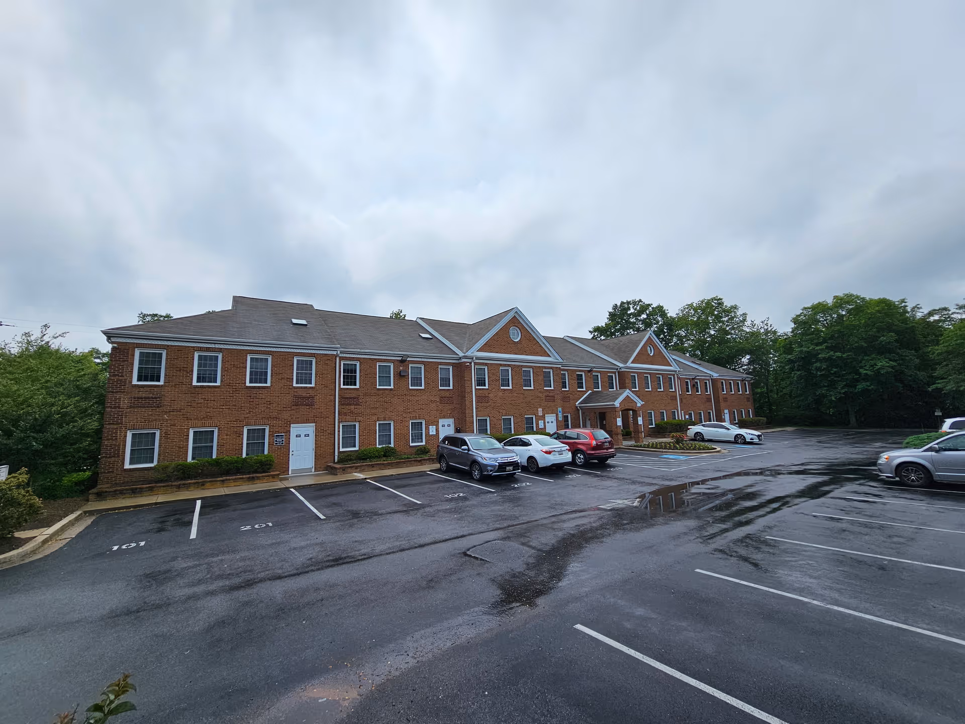 Brick senior living facility with parked cars along a wet driveway and trees nearby