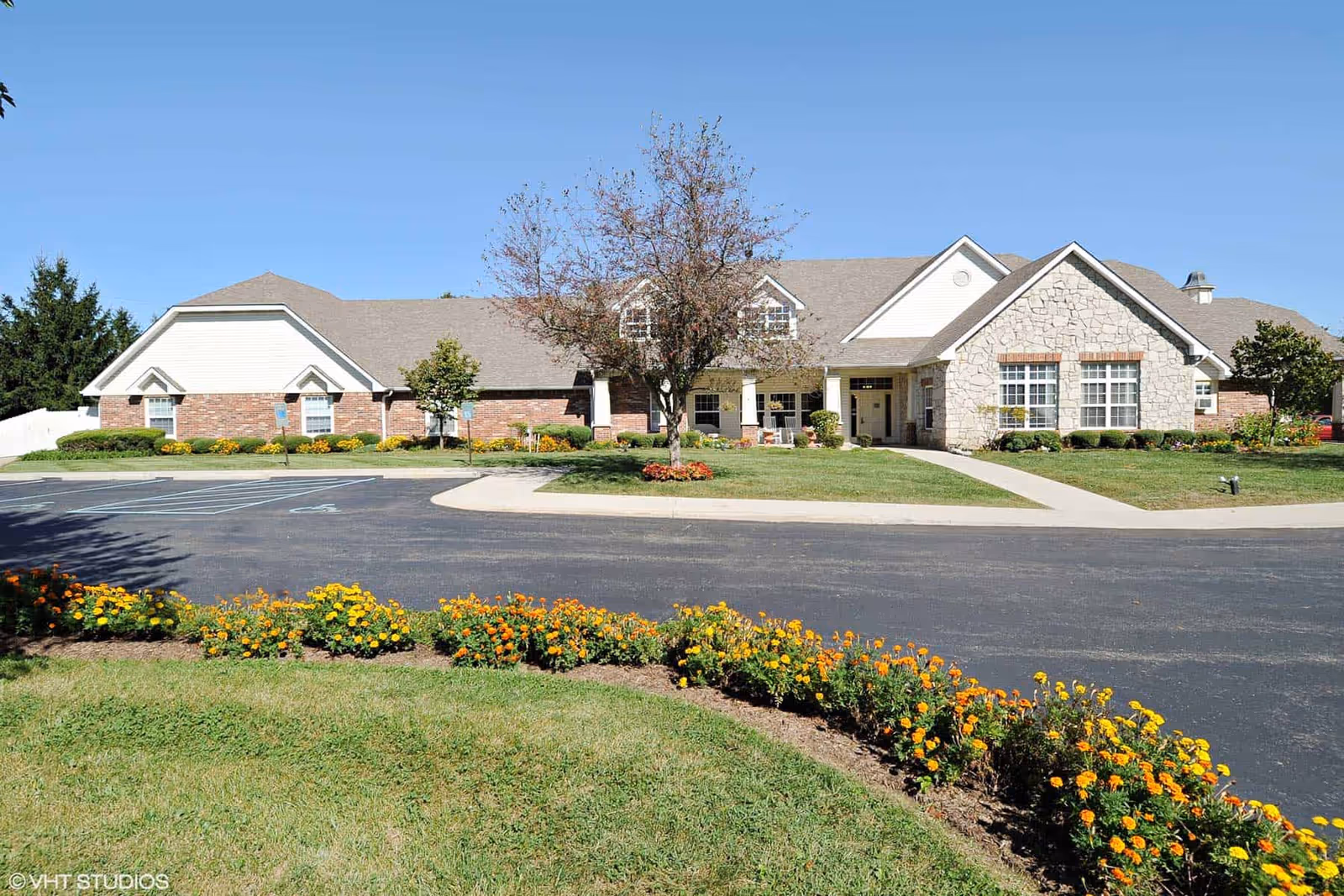 Exterior view of Keepsake Village of Columbus Memory Care building with a paved driveway, landscaped flower beds with orange and yellow flowers, a tree in front, and a clear blue sky.
