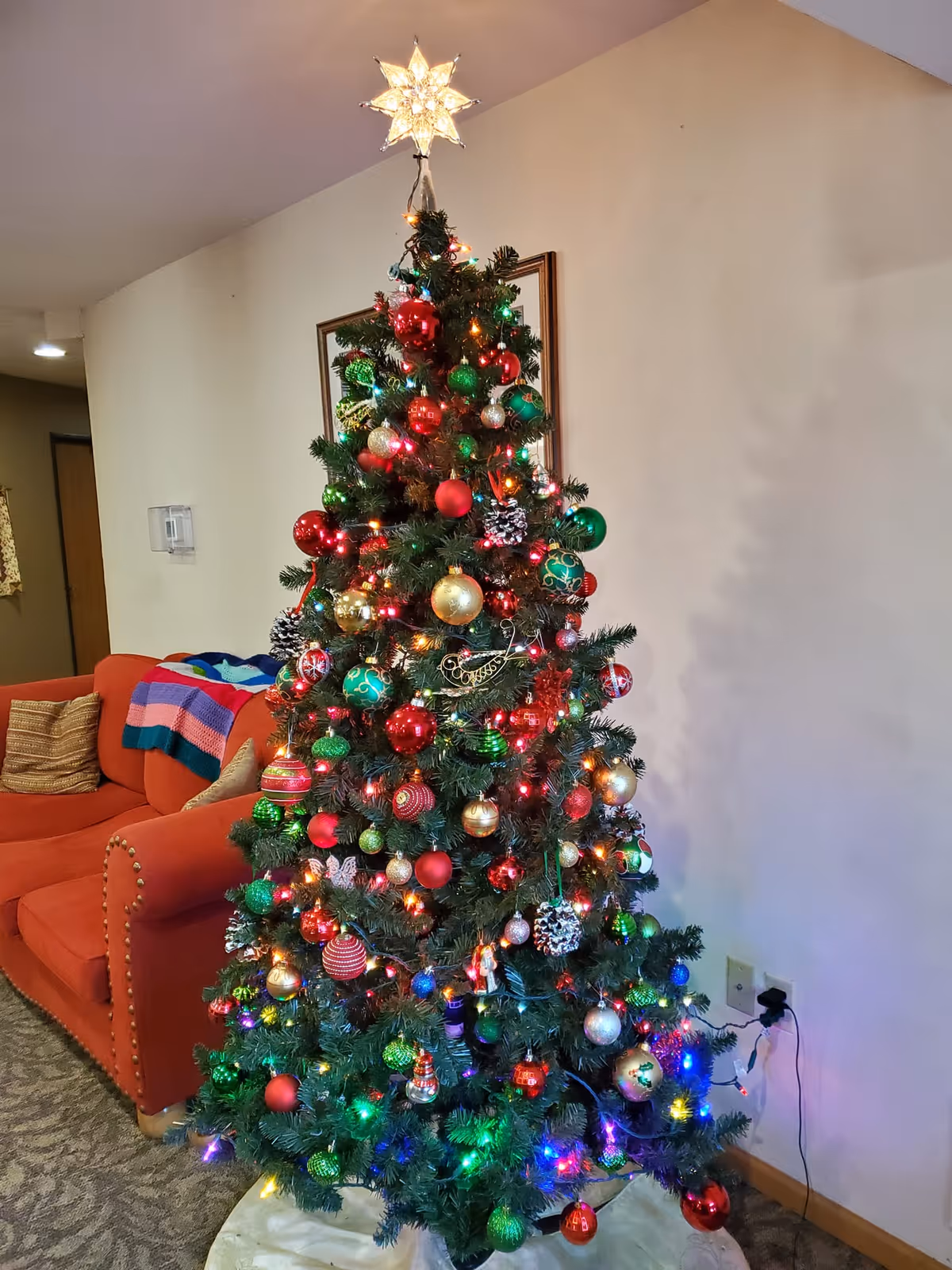 Decorated Christmas tree with multicolored lights and ornaments beside a red couch in a living room.