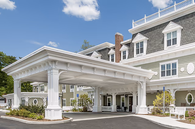 White, columned porte-cochère entrance to a gray-shingled senior living building.
