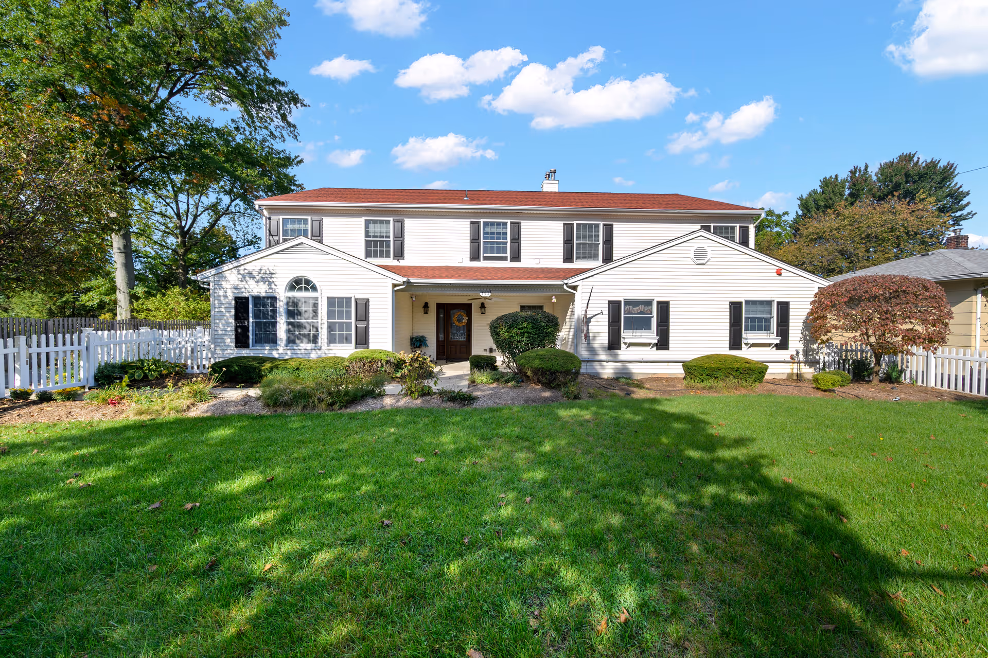 Front view of a two-story white house-like building with a red roof, green lawn, white picket fence, and shrubs.