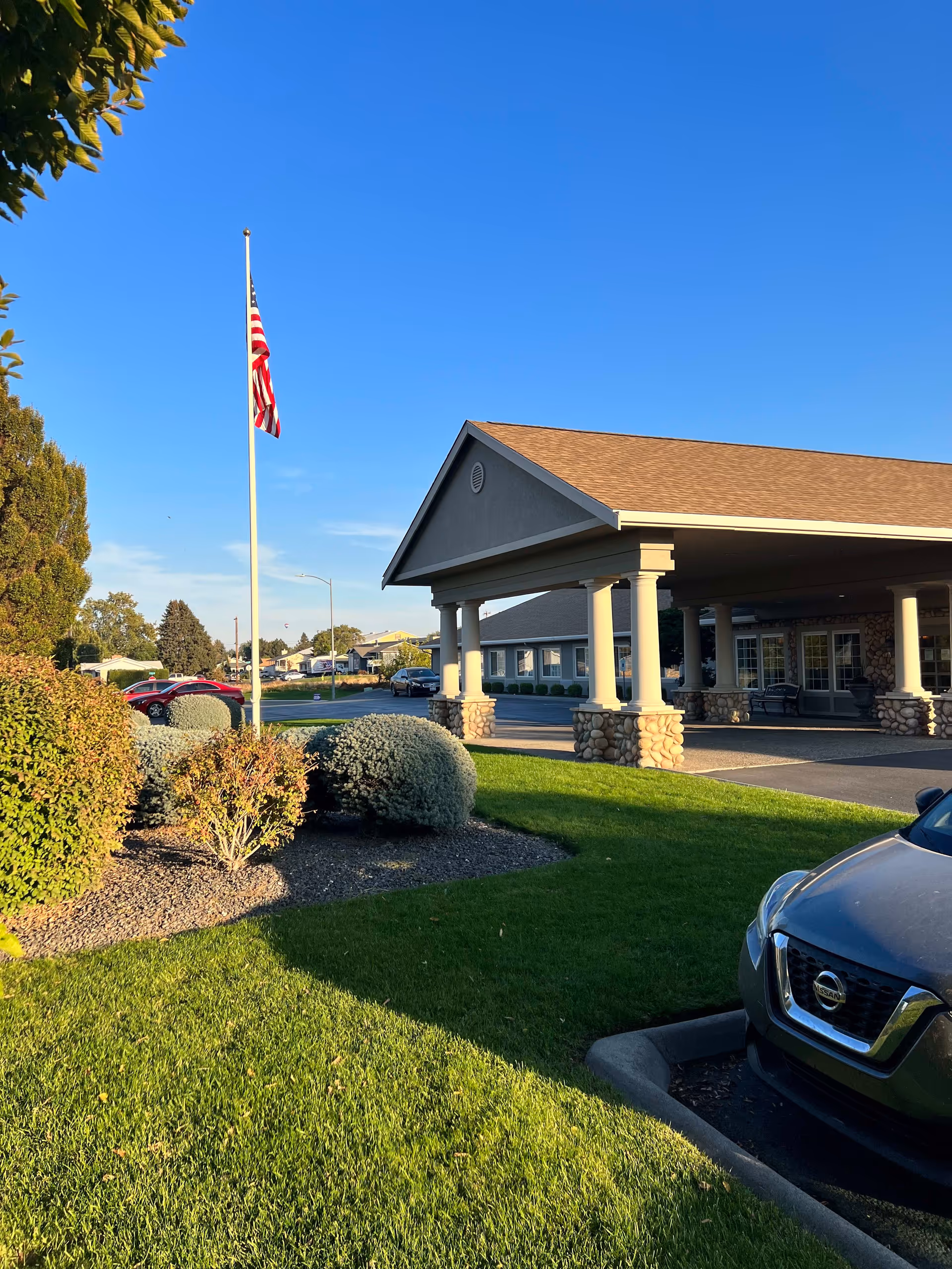 Exterior view of Callaway Gardens Alzheimer's Special Care facility showing a covered entrance with stone pillars, a neatly manicured lawn, bushes, an American flag on a flagpole, and a parked car under clear blue sky.