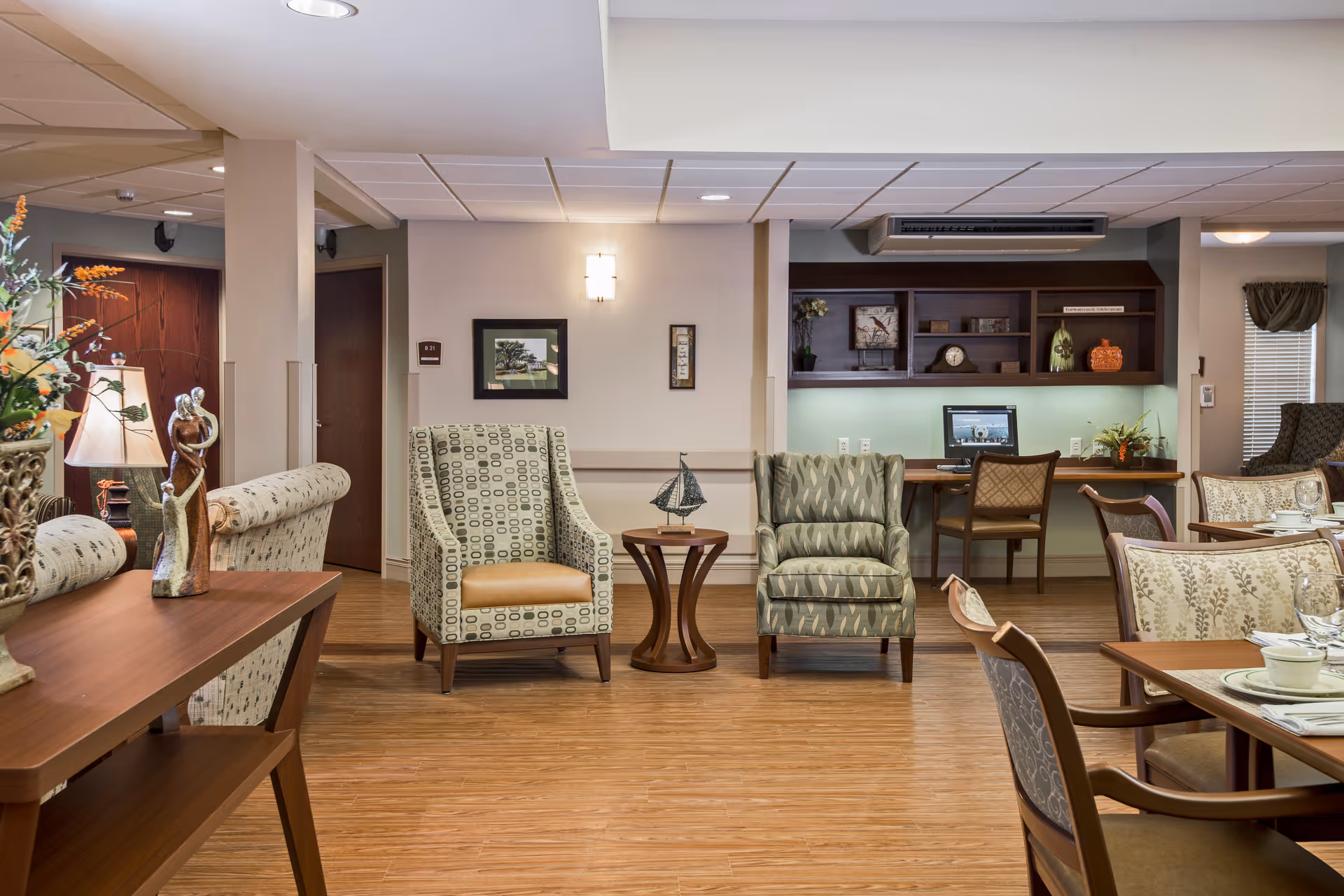 A cozy and well-lit common area in a senior living facility featuring patterned armchairs, a small wooden table with a decorative sailboat, a desk with a computer and chair, and a dining area with tables and chairs set with plates and cups. The room has wooden flooring, soft lighting, and decorative elements including a lamp, sculptures, and framed pictures on the walls.