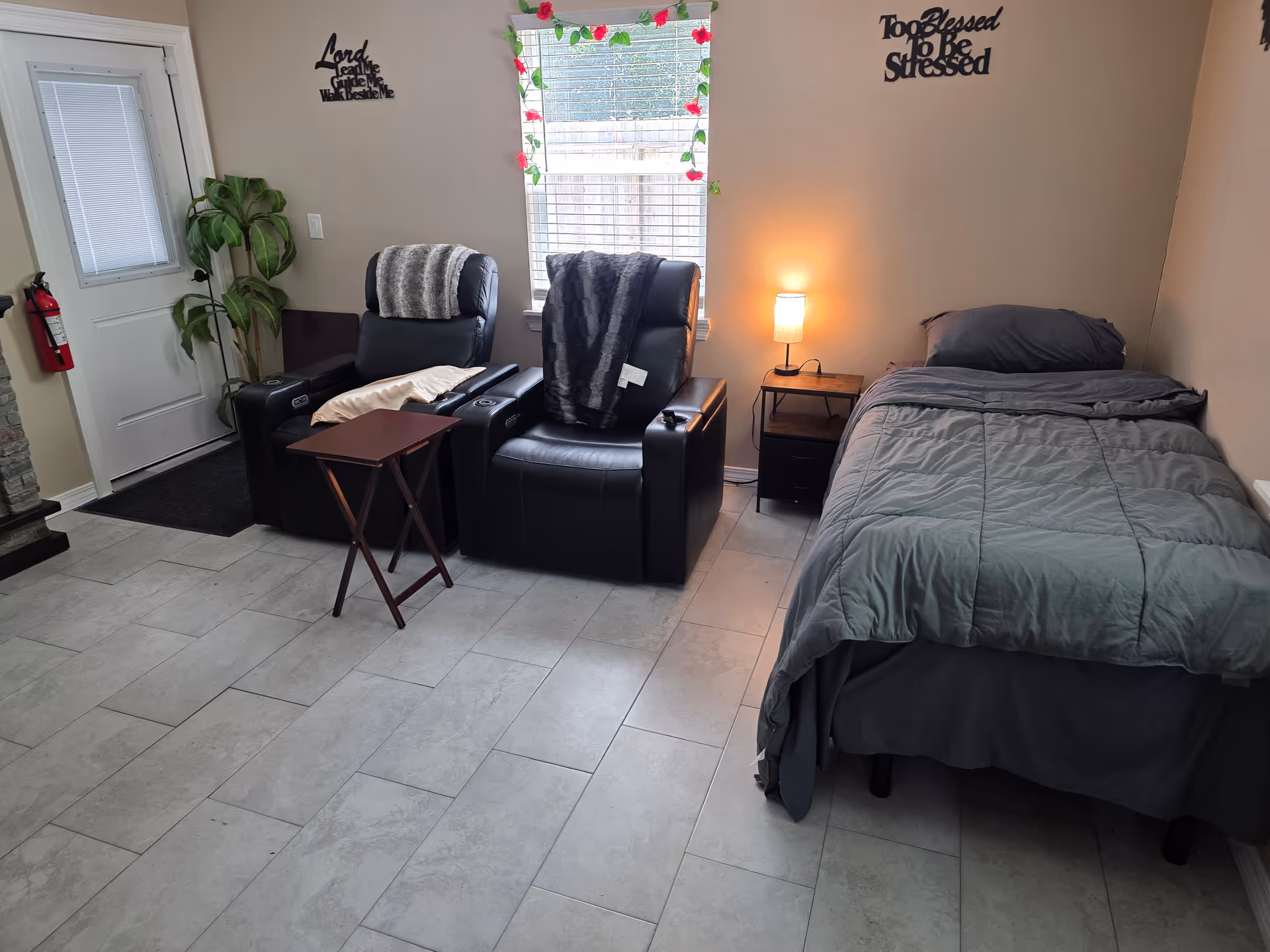 Bedroom in a senior living facility featuring a bed, two recliners, window, and plant decor.
