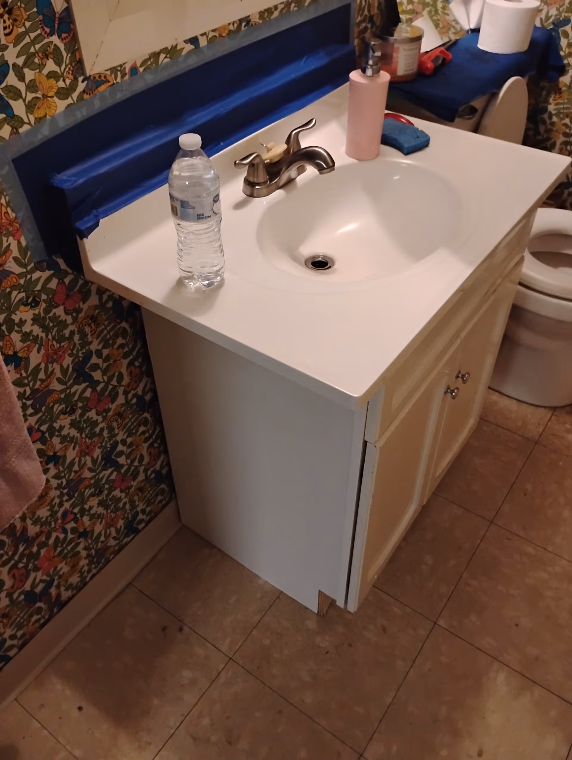 Bathroom vanity with sink, faucet, water bottle, toilet, and floral wallpaper.