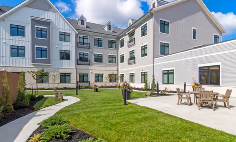 Sunlit courtyard with a green lawn, patio seating, planters, and the multi-story exterior of a senior living building under a blue sky.