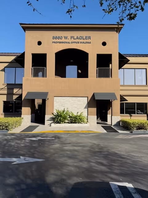Two-story tan office building with arched central opening, balconies, awnings, and landscaped entrance.