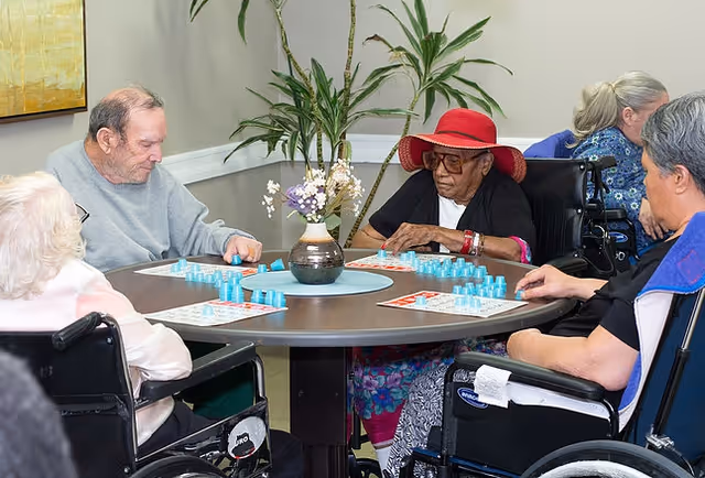 A group of elderly individuals sitting around a round table playing a game with blue pieces. The table has a vase with flowers in the center. Some of the individuals are in wheelchairs, and one woman is wearing a red hat and sunglasses. There are plants and a painting on the wall in the background.