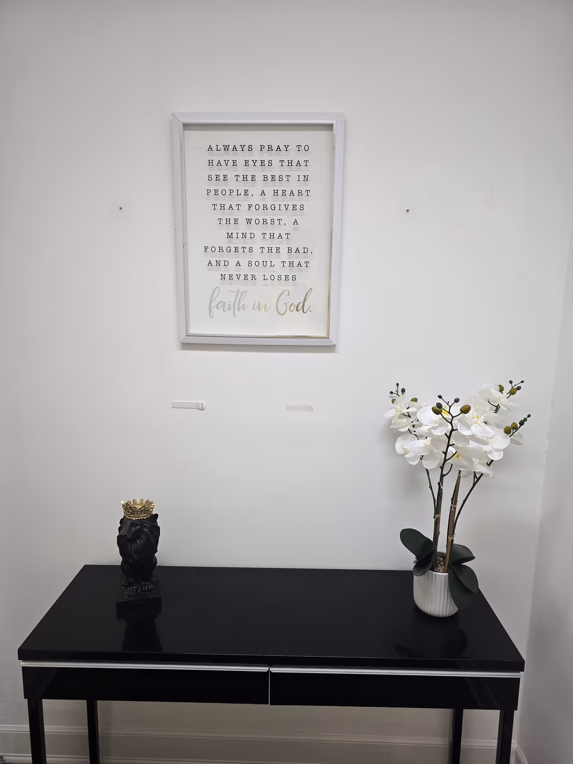 Black glossy console table in a hallway with framed wall art, white orchid, and small statue.