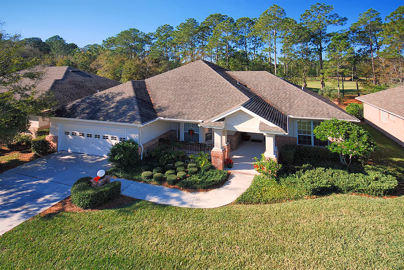 A single-story residential building with a large, sloped roof, a two-car garage, and a covered front porch. The house is surrounded by well-maintained landscaping including bushes, shrubs, and a green lawn. Tall trees are visible in the background under a clear blue sky.