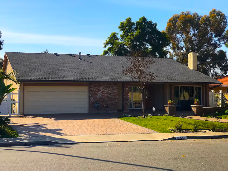 Single-story house with a dark shingled roof, a double garage door, a brick facade section, a small front lawn with a tree, and a paved driveway under a clear blue sky.