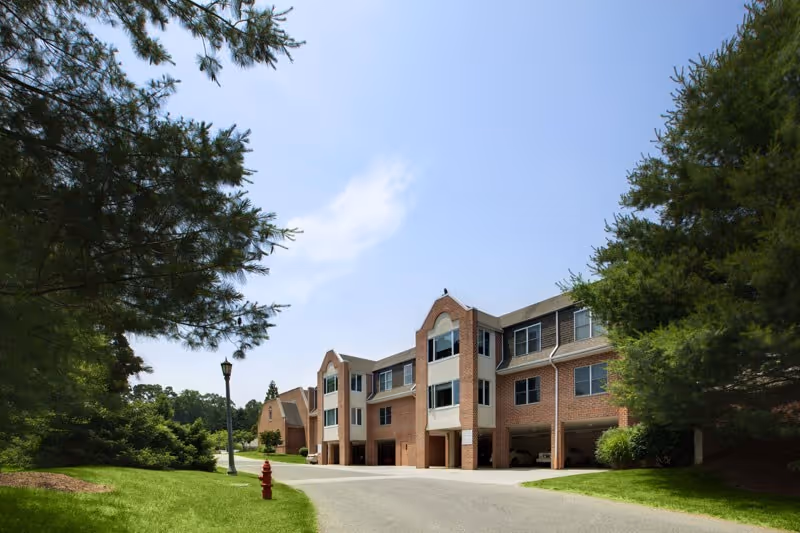 Three-story brick senior living building with covered parking, a paved driveway, and landscaped lawns under a clear sky.