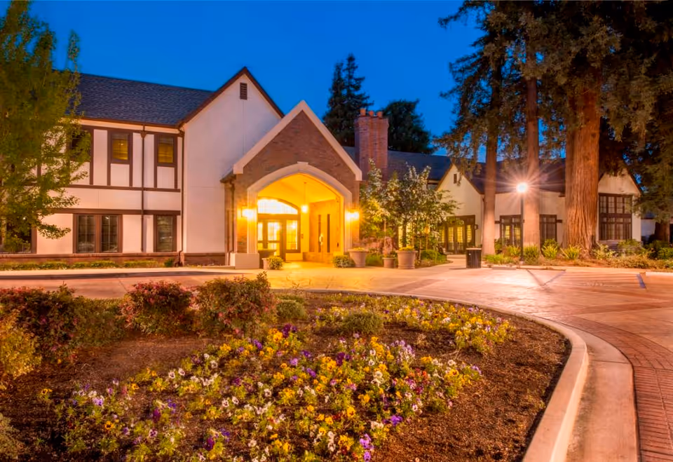 Dusk view of Villa Siena Senior Living Community's illuminated front entrance, Tudor-style building, large trees and a landscaped flower bed.