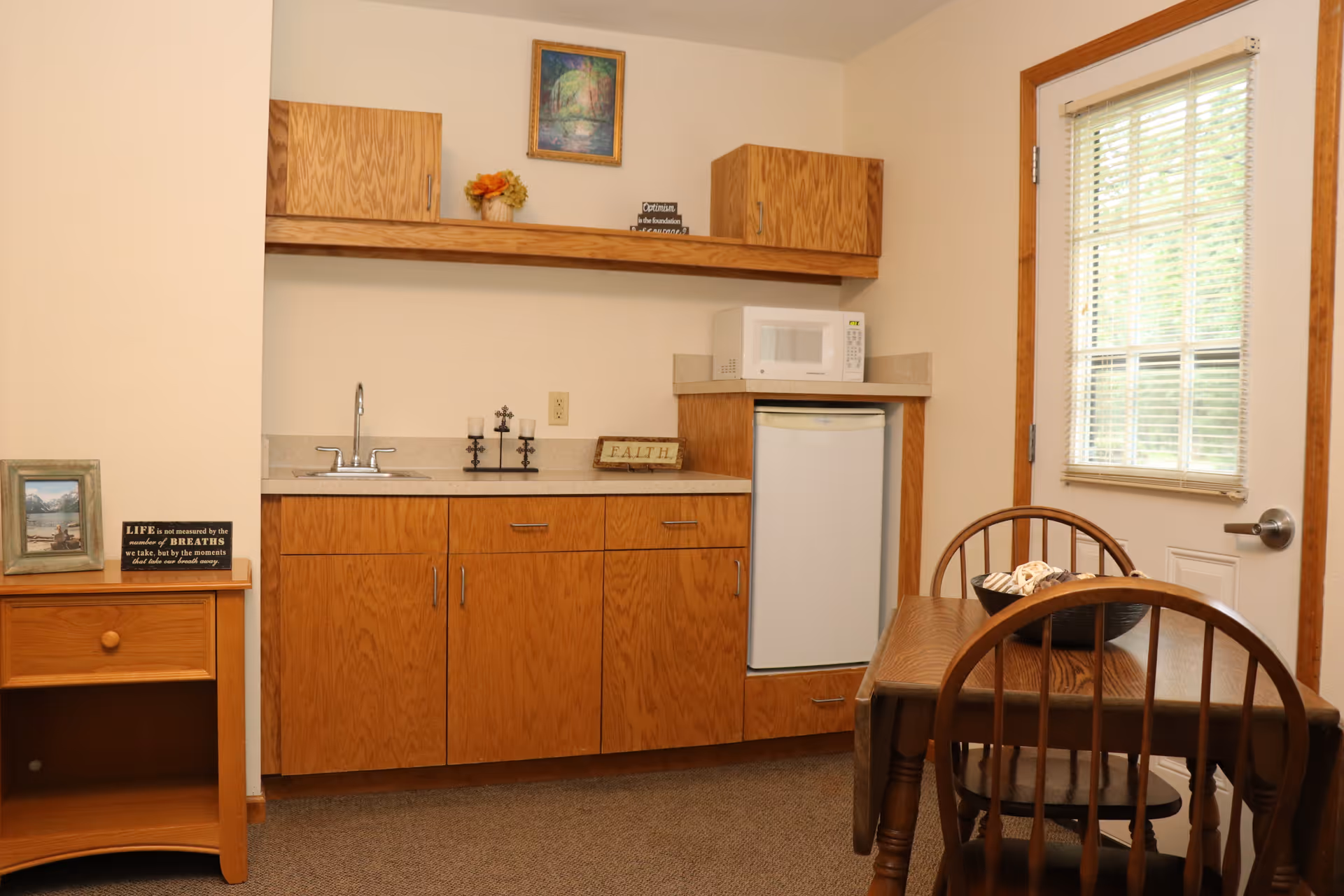 A small kitchenette area with wooden cabinets, a countertop with a sink, a microwave, and a mini refrigerator. There is a wooden table with two chairs next to a door with a window covered by blinds. Decorative items include a framed picture, a small flower arrangement, a sign that says 'FAITH,' and a candle holder on the countertop.