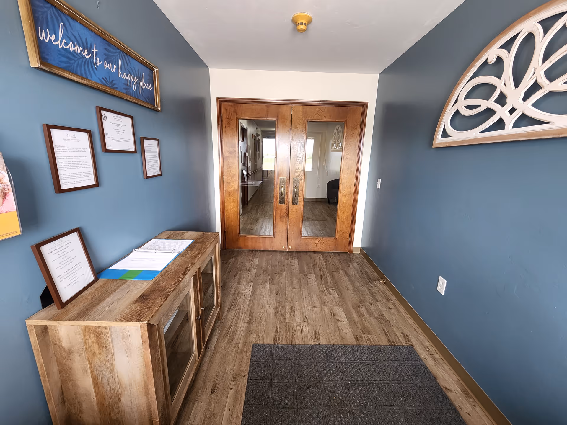 Welcome sign on blue wall beside wooden console and double glass doors.