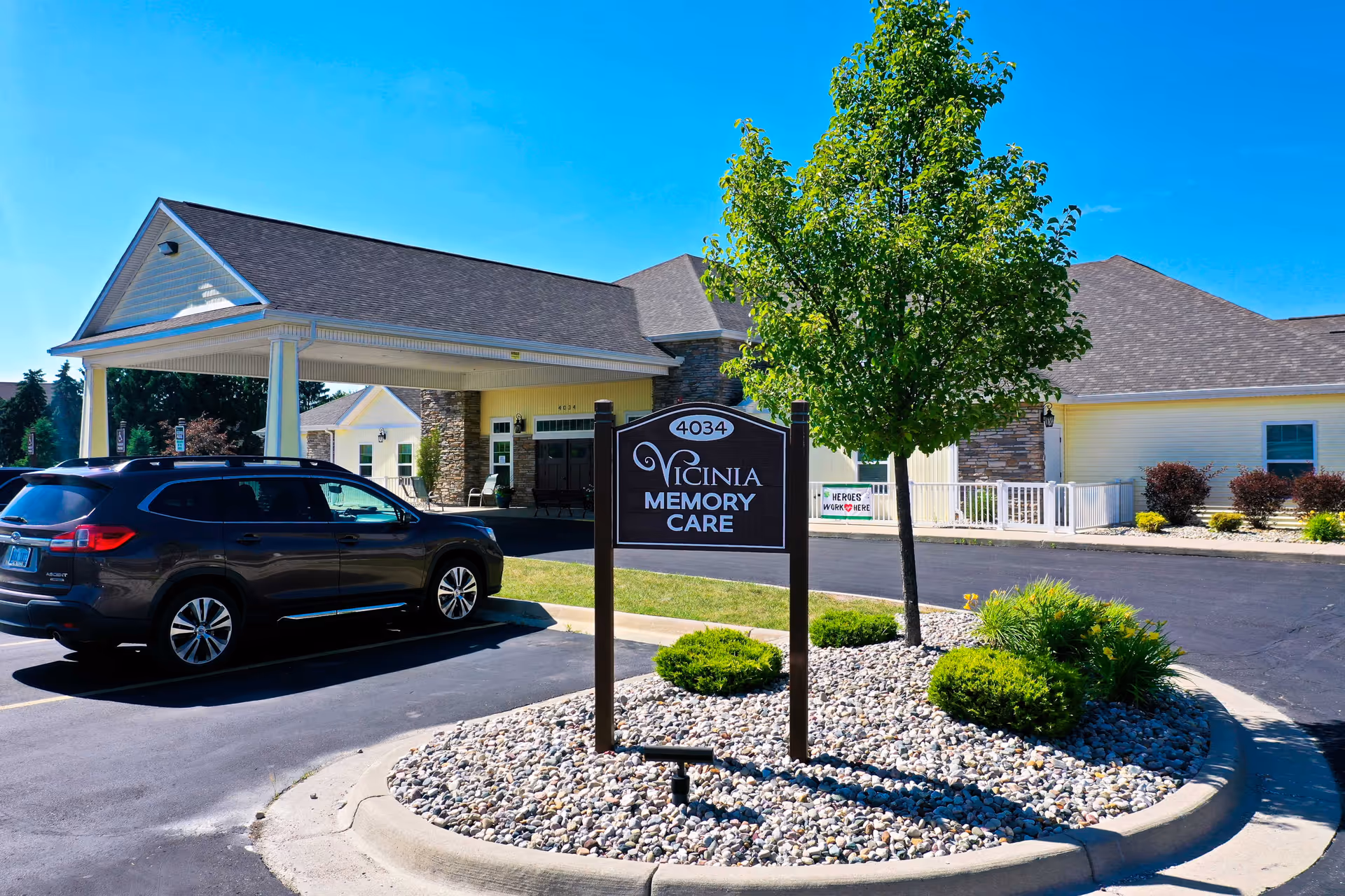 Front entrance of the Vicinia Memory Care facility showing the driveway and parking area with a parked car, entrance canopy, landscaping, and the facility sign under a clear blue sky.