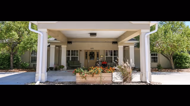 Entrance of a senior living facility named Vintage Park at Holton with a covered porch supported by columns, a planter box with flowers in front, and greenery on either side.