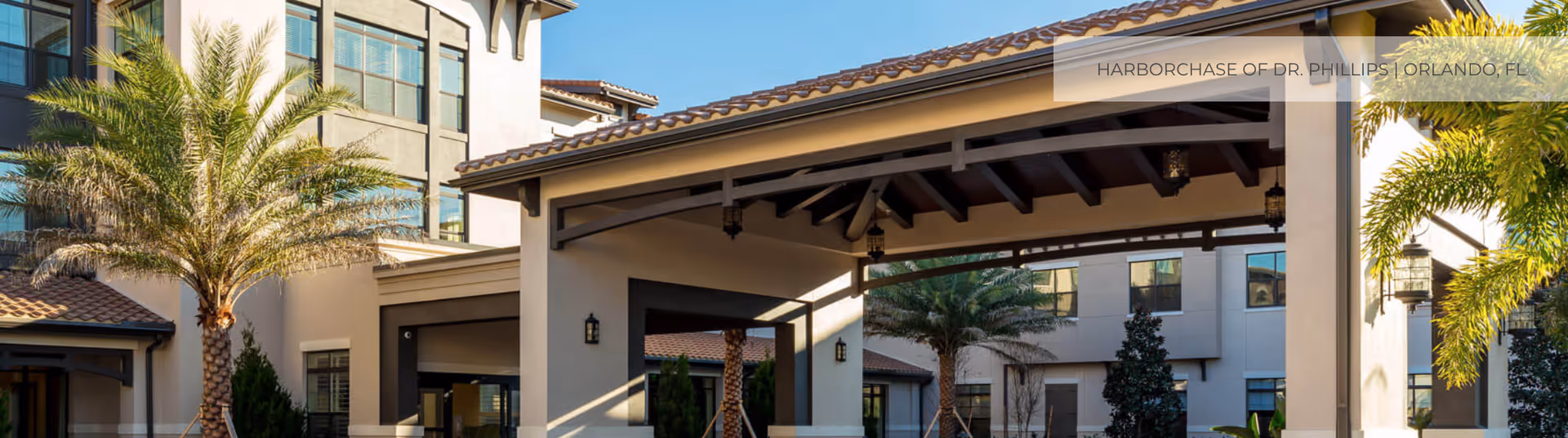 Covered porte-cochère entrance of a senior living building with palm trees and windows in the background.