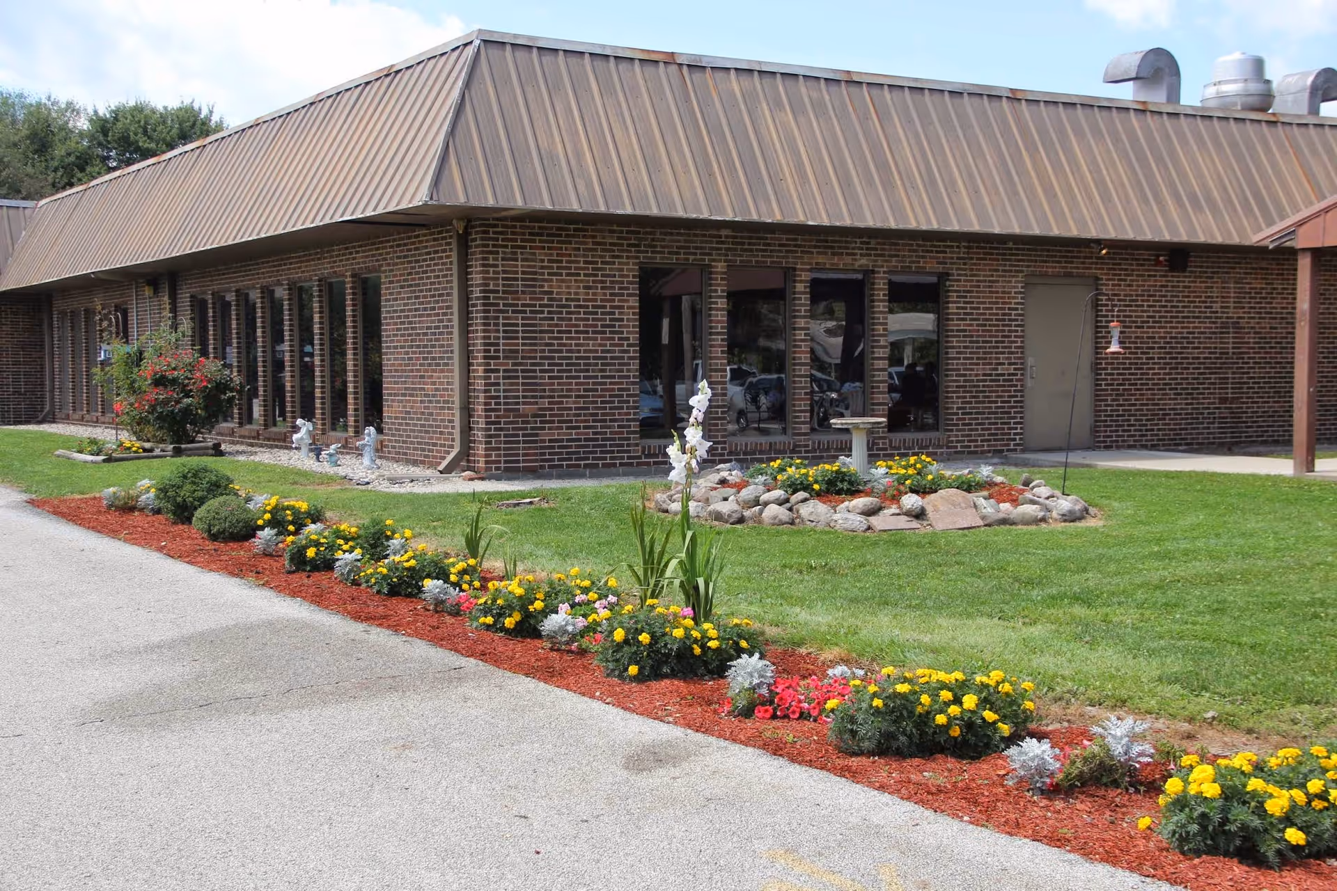 Exterior view of a single-story brick building with a brown metal roof, surrounded by a well-maintained lawn and flower beds with colorful flowers and decorative rocks.