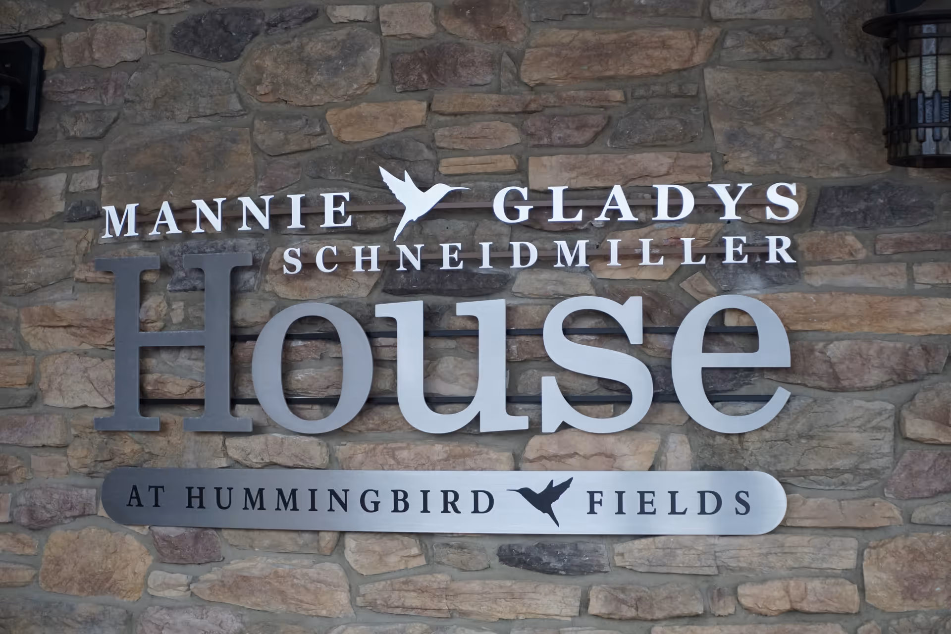 Stone wall sign with large 'House' lettering and names above 'at Hummingbird Fields'.
