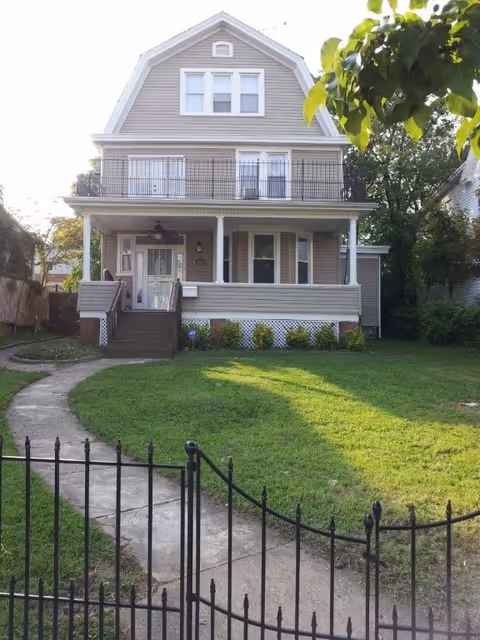 Front exterior of a two-story house with a covered porch, second-floor balcony, curved walkway, and fenced lawn.