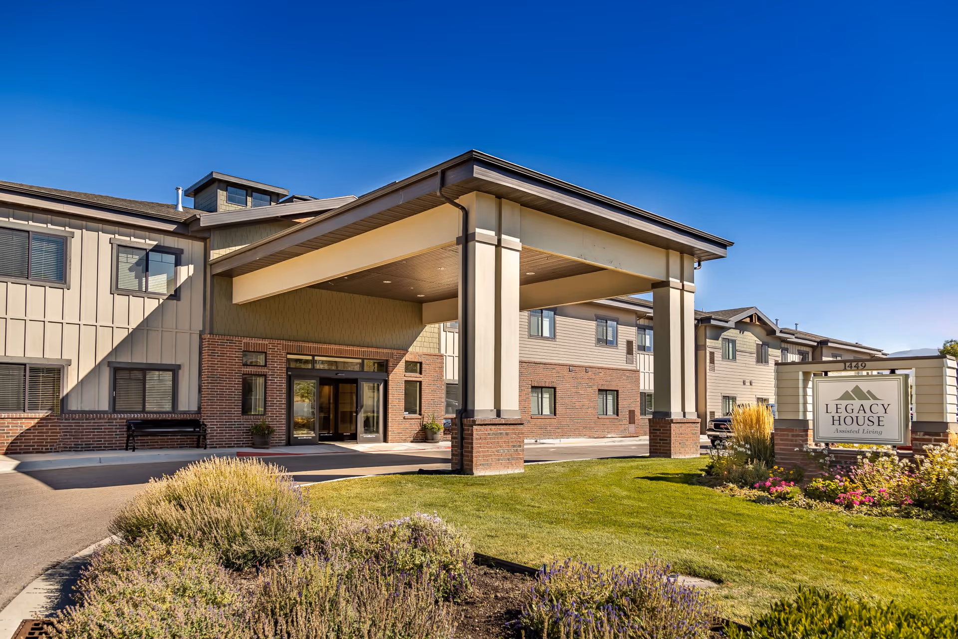 Front entrance of a two-story assisted living building with a covered porte-cochere, landscaping, and a sign reading "Legacy House" under a clear blue sky.