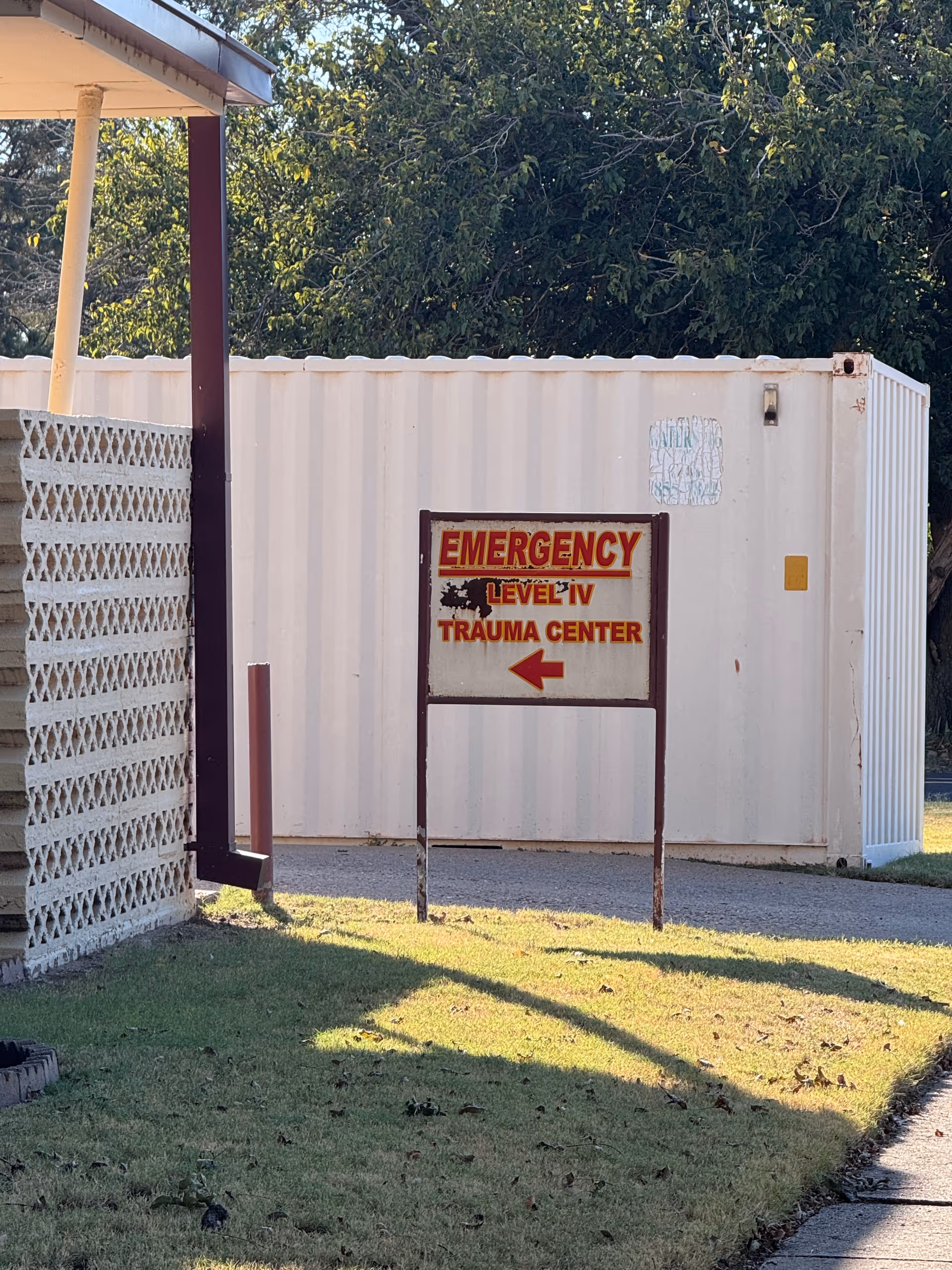 Outdoor scene shows an emergency trauma center sign with arrow beside a white container-like building.