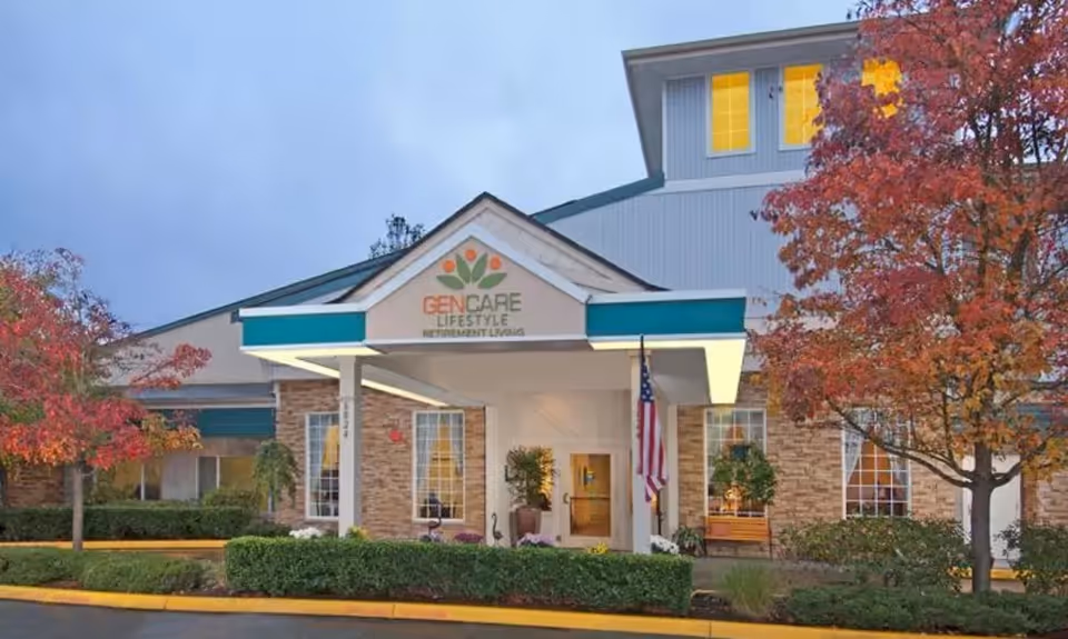 Exterior front view of GenCare Lifestyle Lynnwood at Scriber Gardens building with an entrance canopy, American flag, and autumn trees with red leaves.