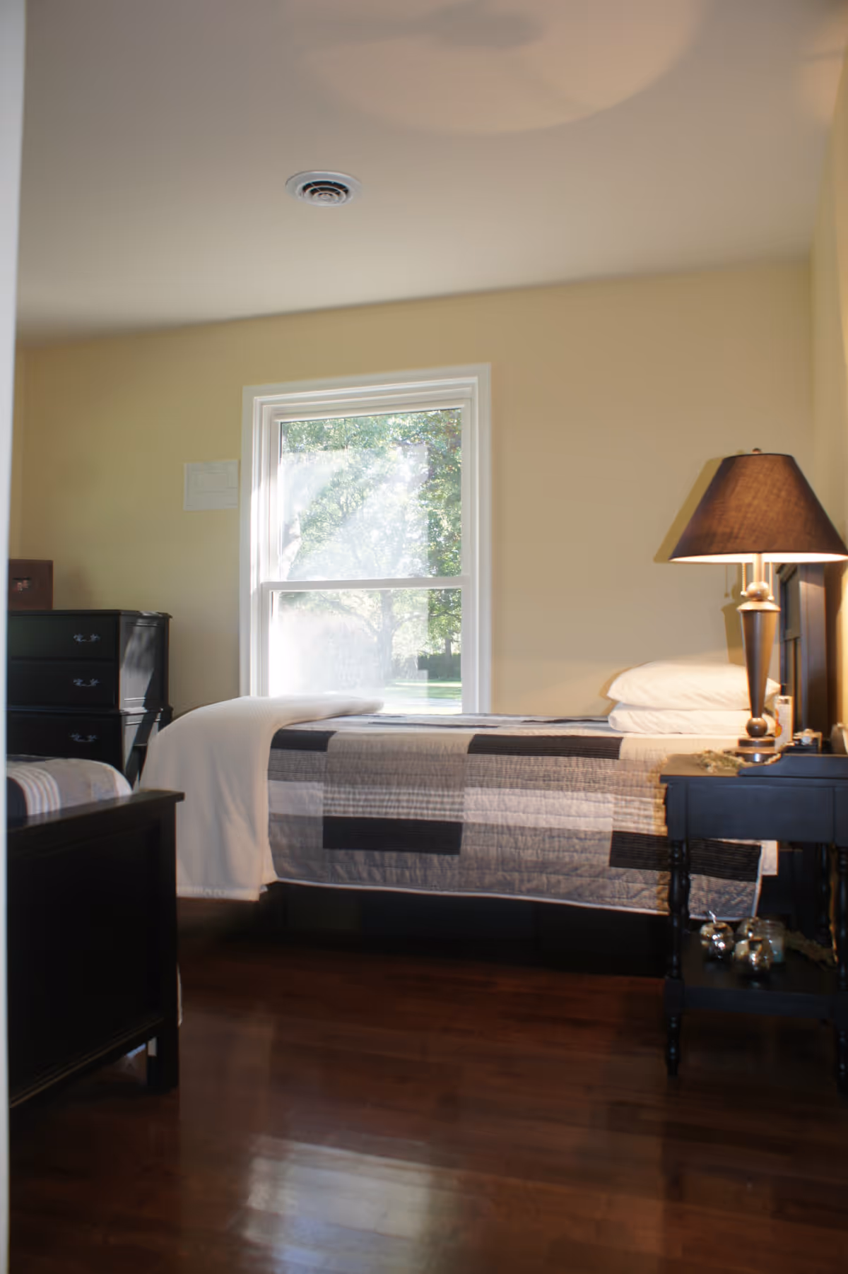 A tidy bedroom with a single bed, patchwork quilt, nightstand, lamp, and dresser.