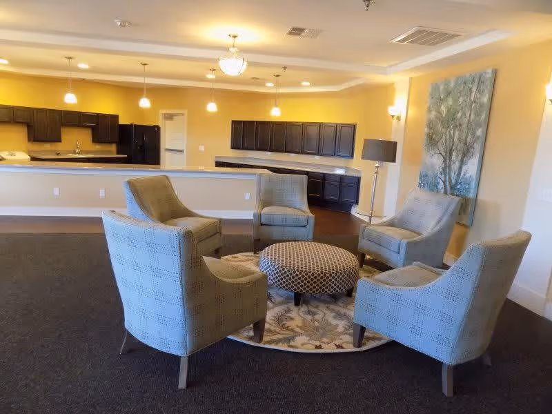A cozy seating area with five upholstered armchairs arranged in a circle around a patterned round ottoman on a floral rug. Behind the seating area is a kitchen with dark wood cabinets, a refrigerator, and a long counter with pendant lights hanging above. The walls are painted a warm yellow, and there is a large painting and a floor lamp on the right side.