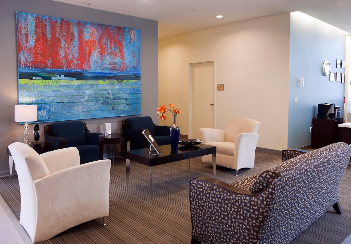 A modern lounge area with a patterned sofa, two beige armchairs, and two dark blue armchairs arranged around a black coffee table with a blue vase holding orange flowers. A large colorful abstract painting hangs on the wall behind the blue chairs, and a lamp is on a small side table. The room has neutral walls and carpeted flooring.