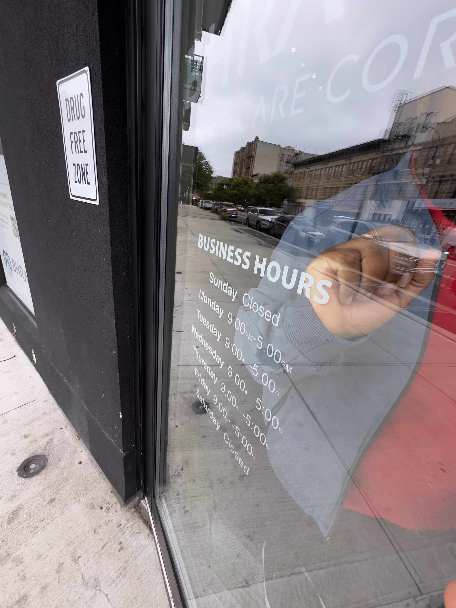Storefront glass displays business hours, street reflections, and a drug-free zone sign.
