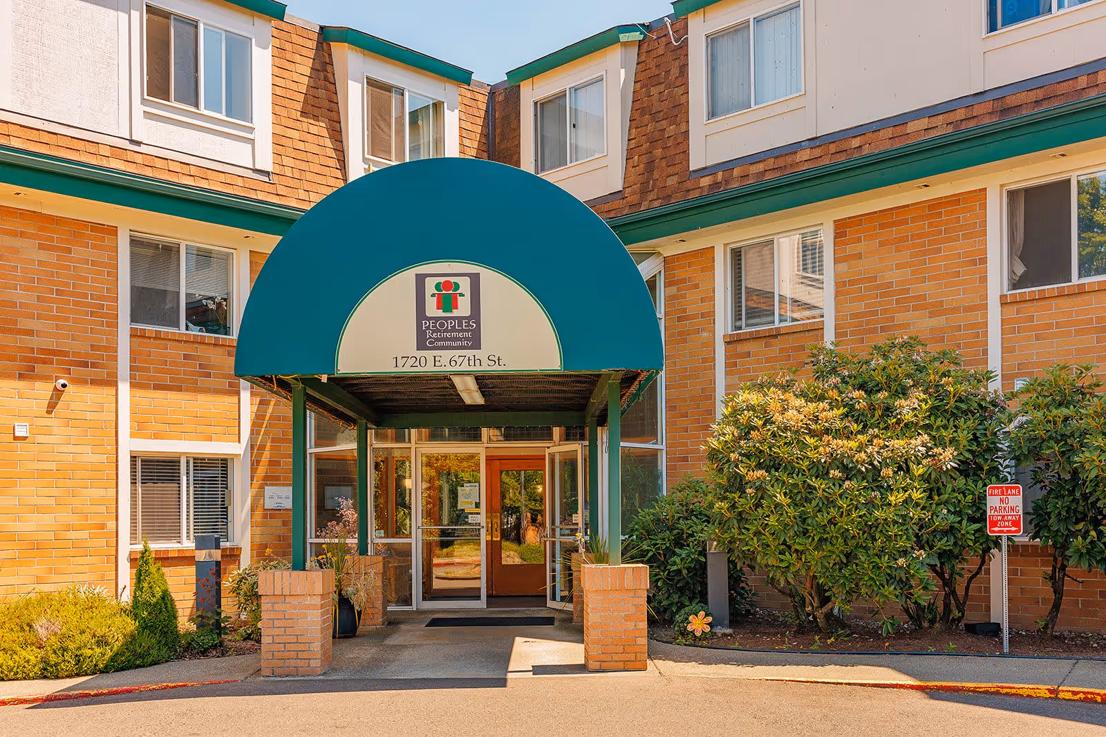 Front entrance of the Peoples Senior Living building with a green arched awning, glass doors, and surrounding brick facade and landscaping.