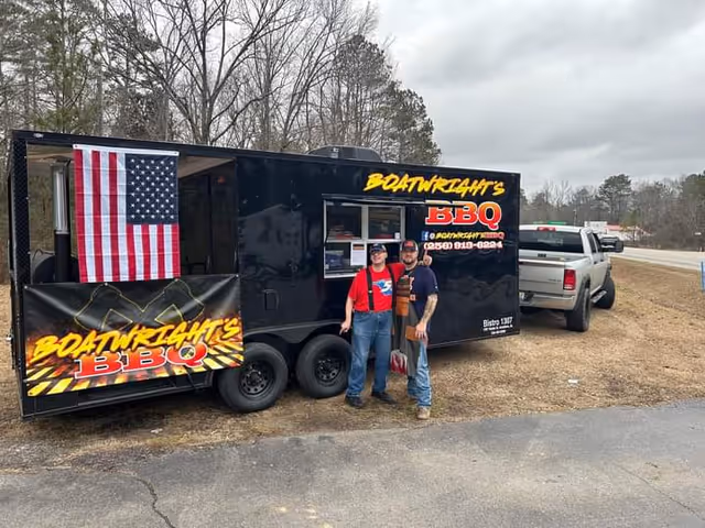 Two men stand beside a black barbecue trailer displaying an American flag.