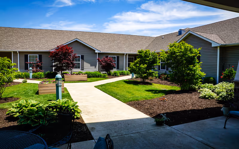 Outdoor courtyard area at Bickford of Crystal Lake featuring a paved walkway, green grass, various shrubs and small trees, and a building with gray siding and multiple windows under a blue sky with some clouds.