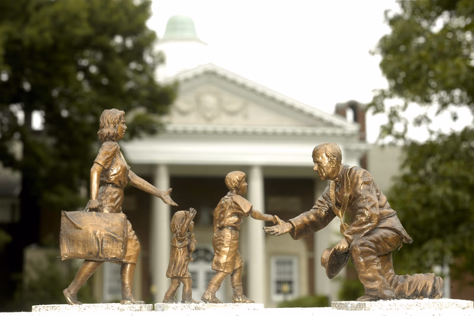 Bronze statues of a woman, child, and man shaking hands before a columned building.