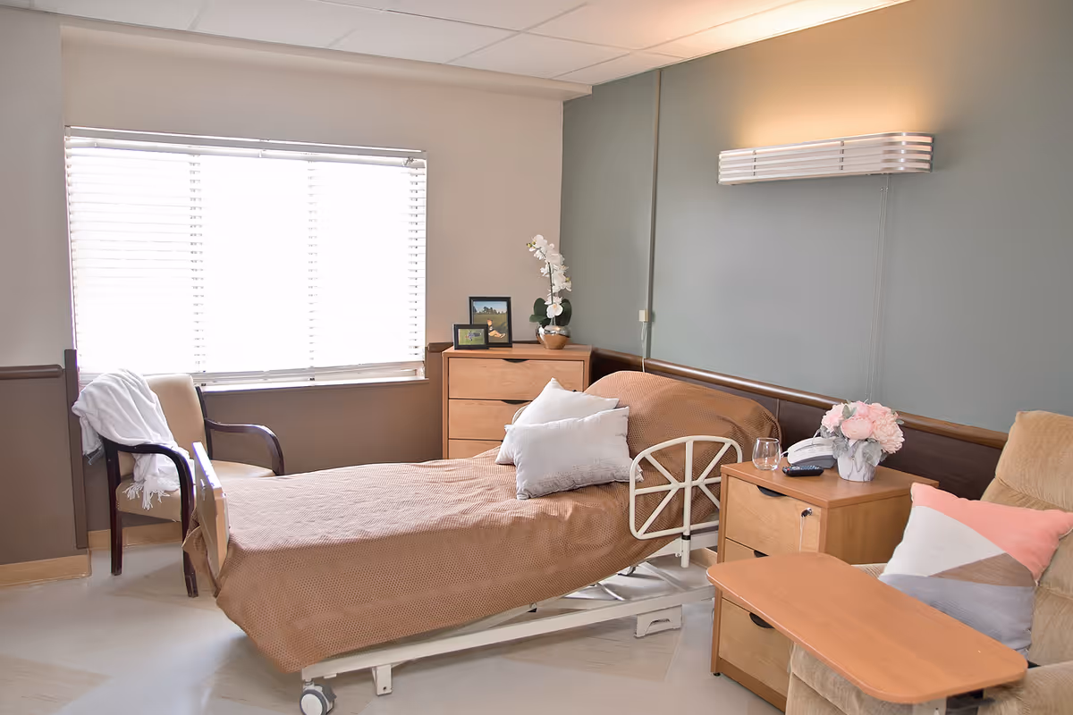 A furnished hospital-style bedroom with a single bed, nightstands, chair, and a window.