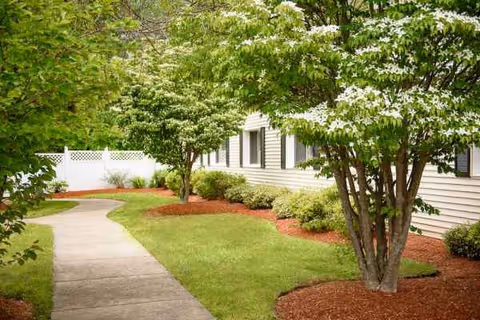 A curved concrete walkway beside a beige senior living building with green trees and shrubs.