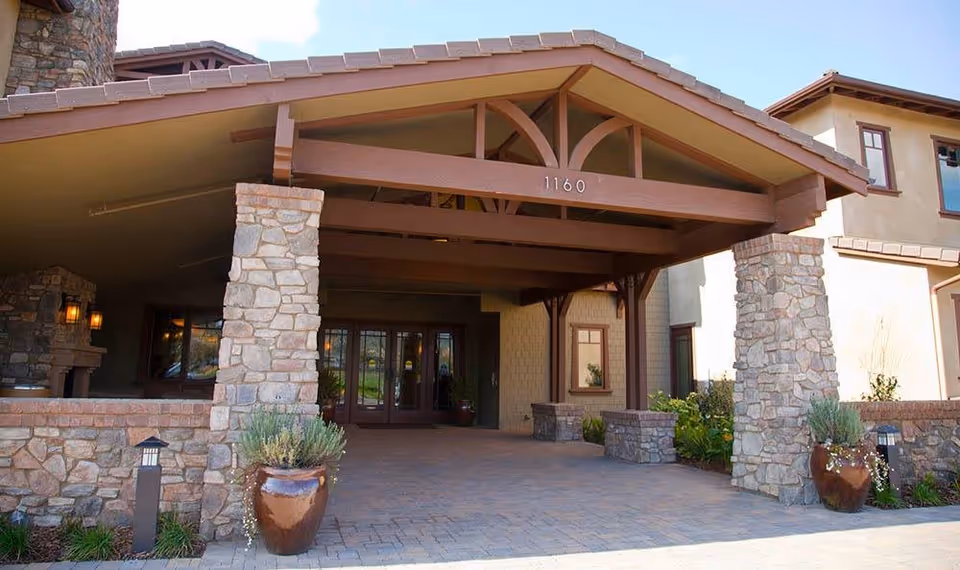 Entrance of a senior living facility with a covered driveway supported by stone pillars and wooden beams. The building has a stone and stucco exterior with large windows and potted plants near the entrance.