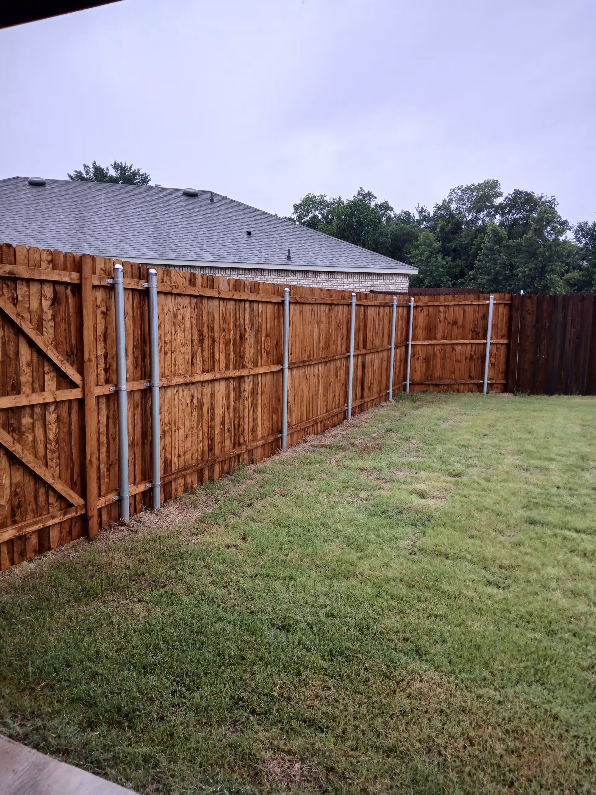 Backyard with wooden fence, green grass, and a house roof in the background.