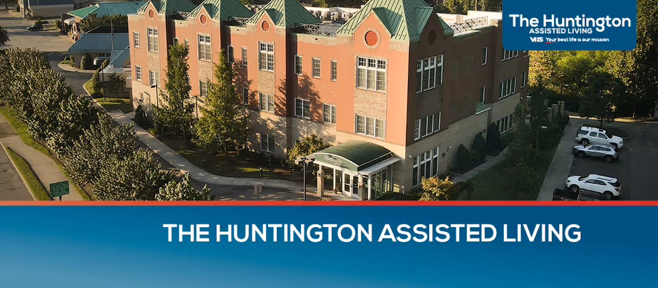 Aerial view of The Huntington Assisted Living building, a multi-story brick structure with green roofs and surrounded by trees and parked cars.