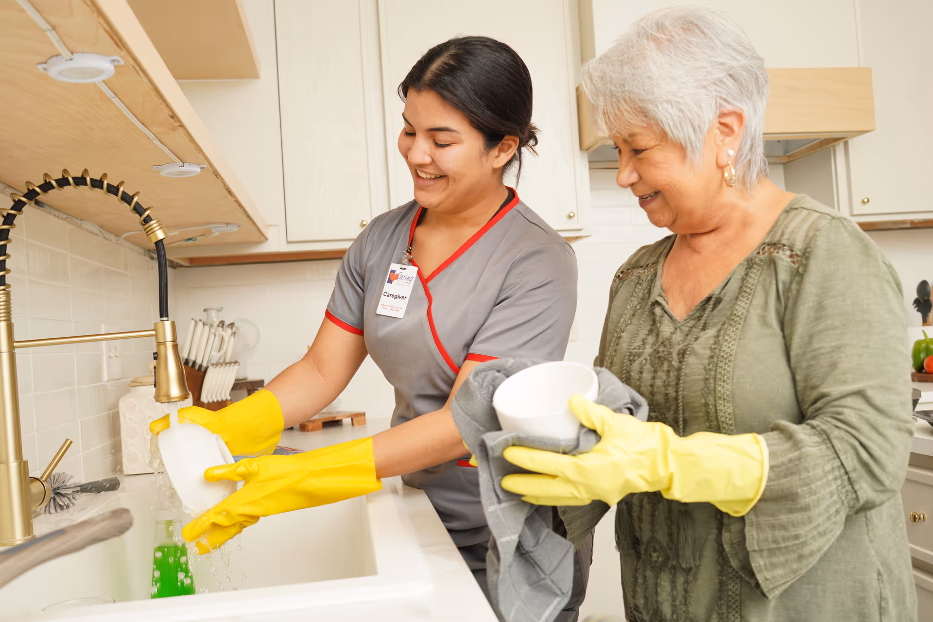 Caregiver helps elderly resident wash dishes at a bright kitchen sink.