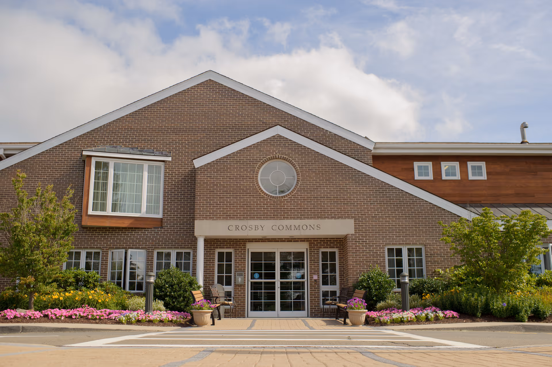 Brick facade and main entrance of Crosby Commons with potted plants, flower beds, benches, and a paved driveway.
