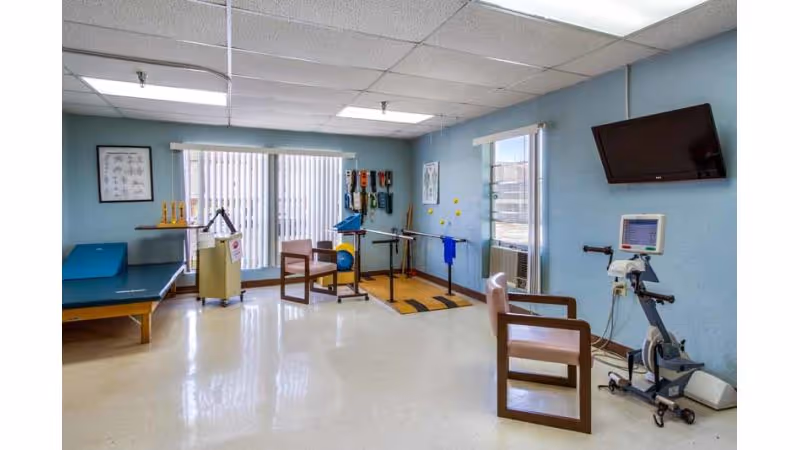 A rehabilitation room with exercise equipment including parallel bars, a stationary bike, chairs, and therapy tools mounted on the wall. The room has light blue walls, large windows with vertical blinds, and a wall-mounted TV.