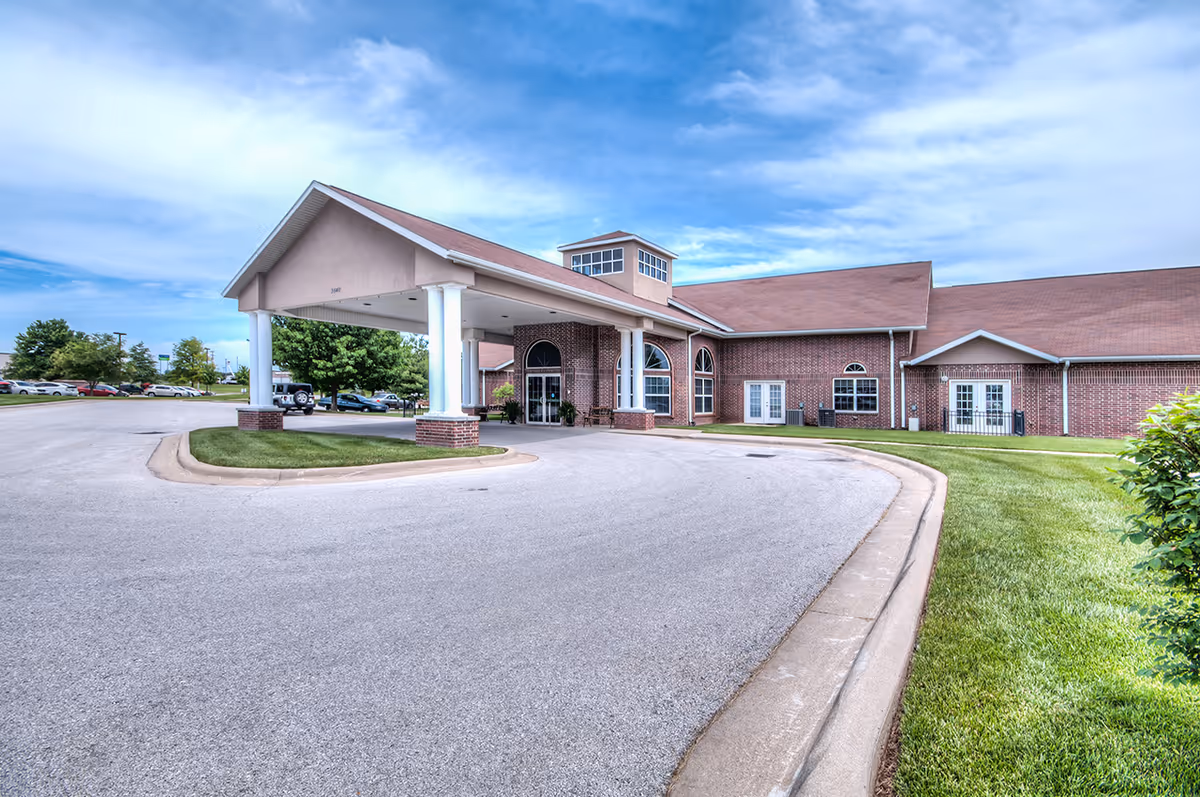 Brick senior living facility with a covered entrance and circular driveway under a blue sky.