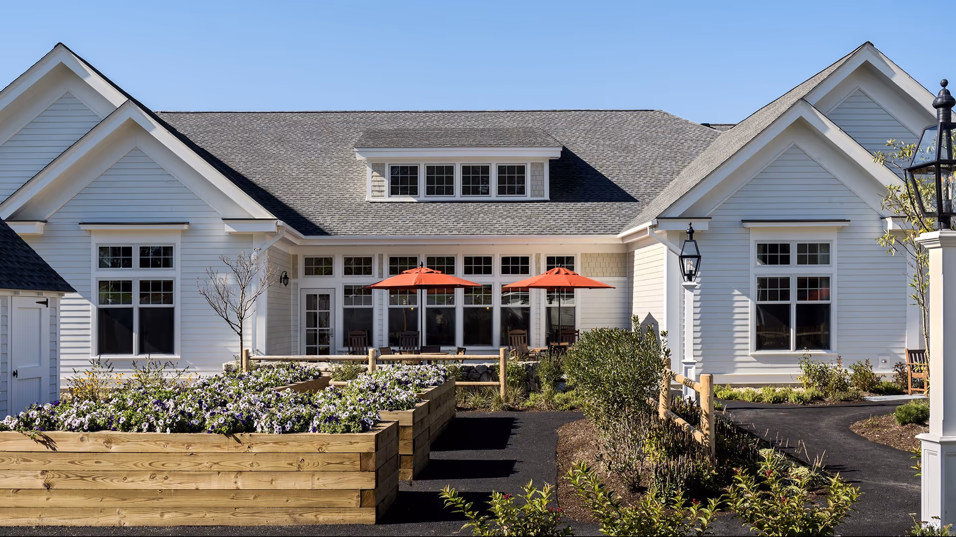 Exterior view of a senior living facility building with white siding, multiple windows, and a gray shingled roof. In front, there are raised wooden garden beds filled with blooming flowers, a paved walkway, and two red patio umbrellas shading outdoor seating areas. The sky is clear and blue.