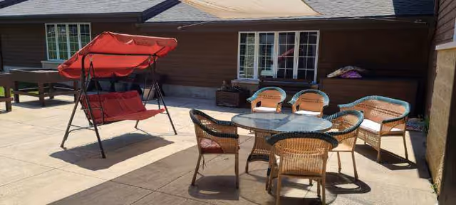 Outdoor patio area with a red cushioned swing, a round glass-top table surrounded by six wicker chairs with cushions, and a foosball table against the building wall under windows.